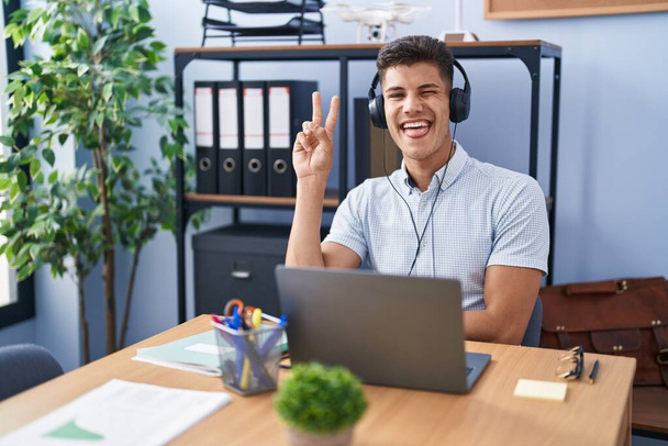 Jeune homme hispanique travaillant au bureau portant des écouteurs souriant avec un sourire heureux clin d'oeil à la caméra faisant signe de victoire. numéro deux.  - Photo, image