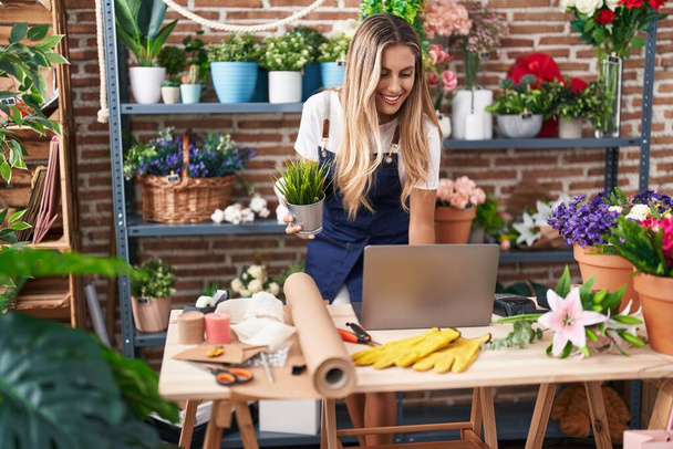 Young blonde woman florist using laptop holding plant at florist - Foto, afbeelding
