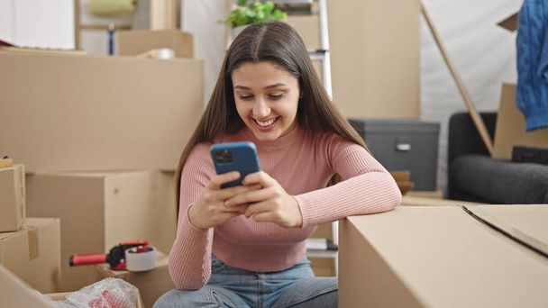 Young beautiful hispanic woman using smartphone sitting on floor at new home - Photo, image