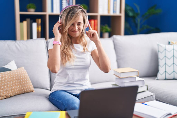 若いブロンドの女性は、大きな音楽のノイズのためにいらいらした表情で指で耳をカバー自宅でコンピュータのラップトップを使用して勉強します。聴覚障害の概念.  - 写真・画像