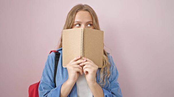 Young blonde woman student covering mouth with book over isolated pink background - Foto, Bild