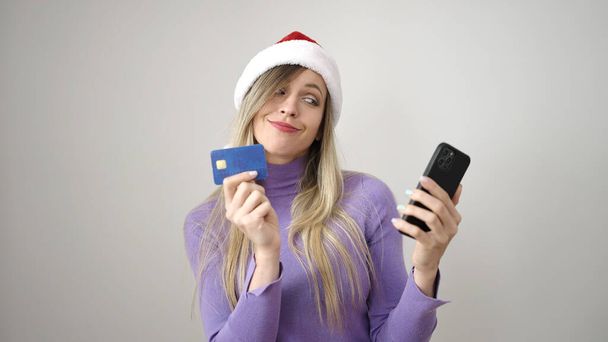 Young blonde woman wearing christmas hat using smartphone and credit card over isolated white background - Zdjęcie, obraz