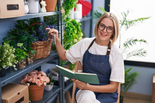 Young blonde woman florist smiling confident reading notebook at florist shop - 写真・画像