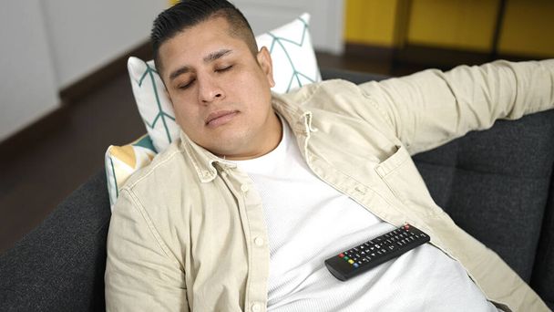 Joven hispano sosteniendo control remoto de televisión acostado en sofá durmiendo en casa - Foto, imagen