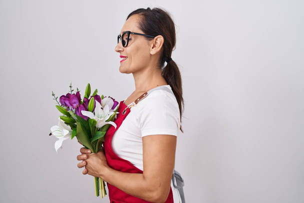 中年ブルネット女性身に着けていますエプロン作業で花屋の店を保持花束側に見て、自然な顔と自信を持って笑顔とプロファイルのポーズをリラックス.  - 写真・画像