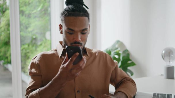 Attentive freelancer listening smartphone voice message indoors closeup. Modern guy using cellphone sitting table alone. African american person sticks to ear loudspeaker looking phone screen at home - Foto, Bild
