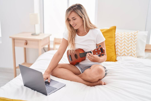 Giovane donna bionda che ha lezione di ukulele online seduta sul letto in camera da letto - Foto, immagini