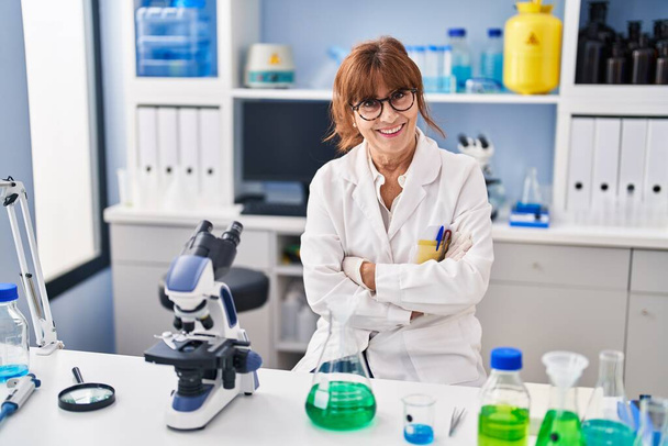 Orta yaşlı kadın bilim adamı gülümsüyor. Kollarını kavuşturup laboratuvarda oturuyor. - Fotoğraf, Görsel