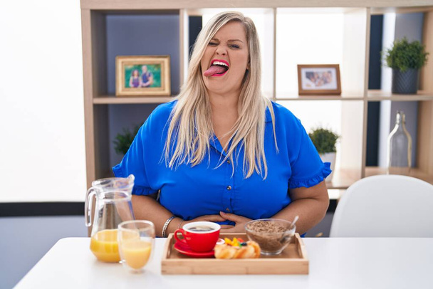 Caucasian plus size woman eating breakfast at home sticking tongue out happy with funny expression. emotion concept.  - 写真・画像