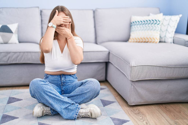 Молода біла жінка сидить на підлозі у вітальні, покриваючи очі і рот руками, здивована і шокована. приховування емоцій
  - Фото, зображення