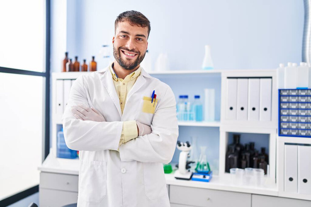 Young hispanic man wearing scientist uniform standing with arms crossed gesture at laboratory - Foto, afbeelding
