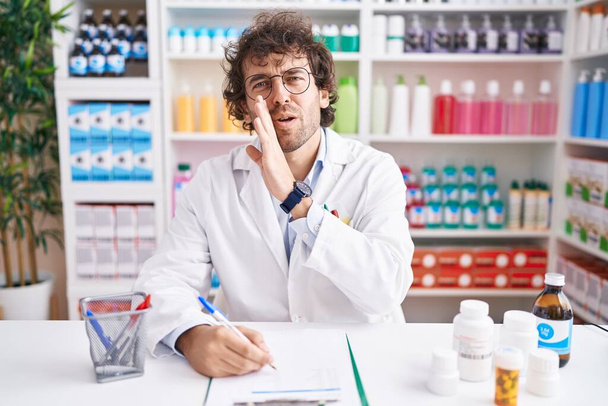 Hispanic young man working at pharmacy drugstore hand on mouth telling secret rumor, whispering malicious talk conversation  - Fotó, kép