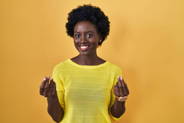 Afričanky mladá žena stojící nad žluté studio dělat peníze gesta s rukama, žádat o výplatu mzdy, milionář podnikání  - Fotografie, Obrázek