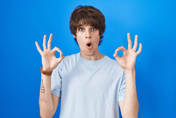 Hispanic young man standing over blue background looking surprised and shocked doing ok approval symbol with fingers. crazy expression  - Zdjęcie, obraz