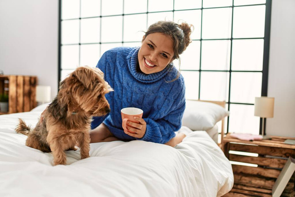 Junge schöne hispanische Frau trinkt eine Tasse Kaffee im Bett mit Hund im Schlafzimmer - Foto, Bild
