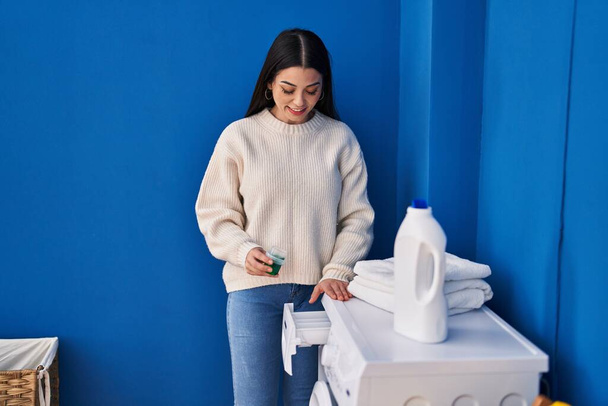 Joven hermosa mujer hispana sonriendo con confianza vertiendo detergente en la lavadora en la sala de lavandería - Foto, Imagen