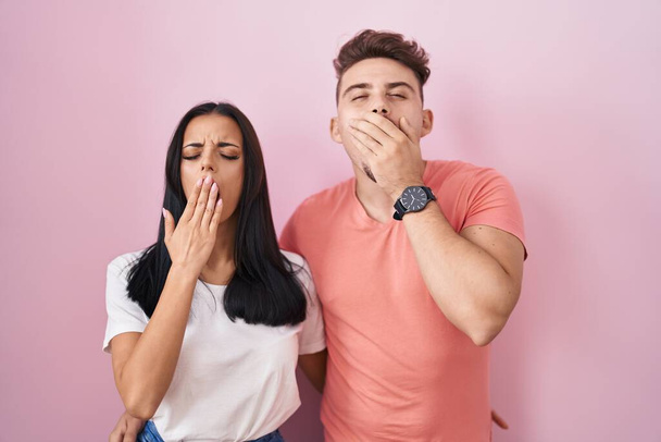 Молодая испаноязычная пара стоящая на розовом фоне скучает зевая усталая закрывая рот рукой. беспокойство и сонливость.  - Фото, изображение