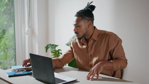 Focused man taking glasses on in office closeup. Dreadlocks guy working laptop at home remotely. Serious freelancer looking computer screen preparing taking notes. Afro american student sitting alone  - Foto, imagen