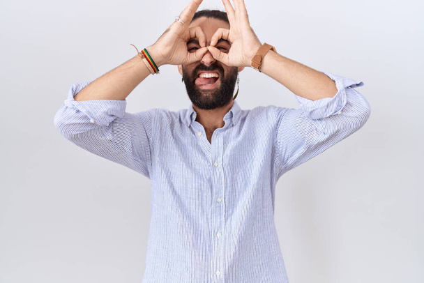 Homme hispanique avec barbe portant une chemise décontractée faisant un geste correct comme des jumelles sortant la langue, les yeux regardant à travers les doigts. expression folle.  - Photo, image