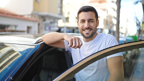 Joven hombre hispano sonriendo confiado apoyado en la puerta del coche en la calle - Foto, Imagen
