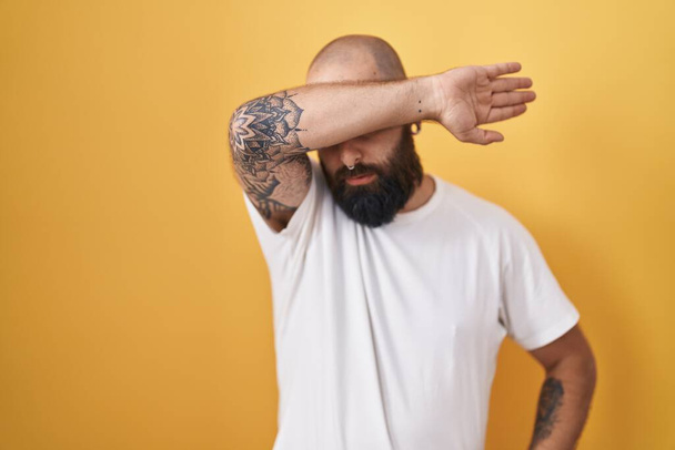 Young hispanic man with beard and tattoos standing over yellow background covering eyes with arm, looking serious and sad. sightless, hiding and rejection concept  - Fotoğraf, Görsel