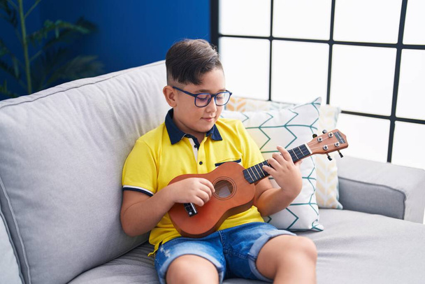 Adorable hispanic boy playing ukulele sitting on sofa at home - 写真・画像