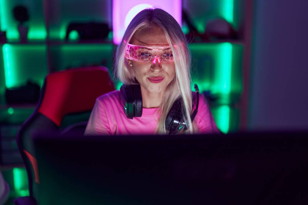 Νεαρή ξανθιά γυναίκα streamer χρησιμοποιώντας υπολογιστή και γυαλιά εικονικής πραγματικότητας στο gaming room - Φωτογραφία, εικόνα