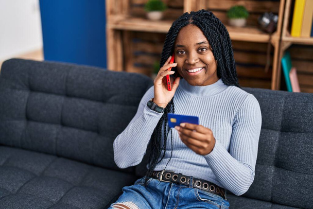 Mujer afroamericana hablando en smartphone usando tarjeta de crédito sentada en sofá en casa - Foto, imagen