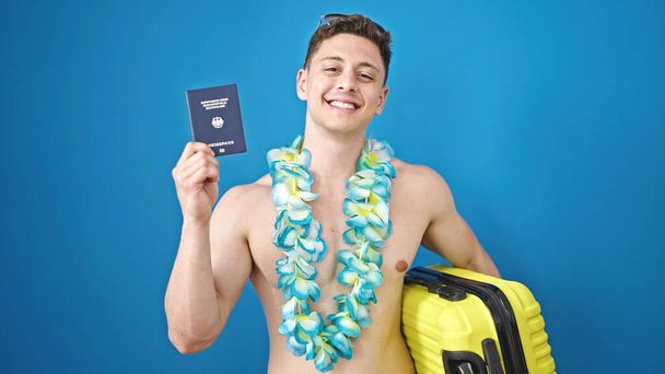 Νεαρός Ισπανός τουρίστας που κρατάει διαβατήριο και βαλίτσα πάνω από απομονωμένο μπλε φόντο - Φωτογραφία, εικόνα