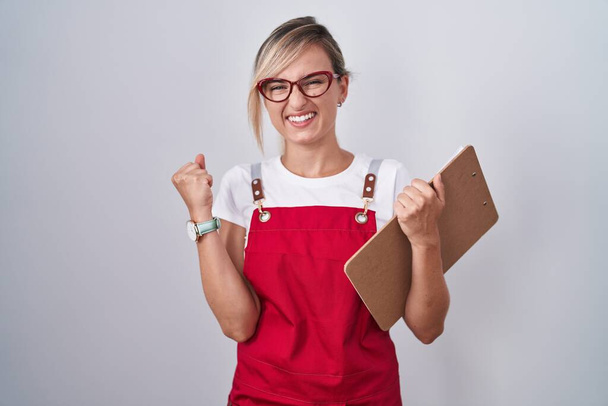 Young blonde woman wearing waiter uniform holding clipboard very happy and excited doing winner gesture with arms raised, smiling and screaming for success. celebration concept.  - Fotografie, Obrázek