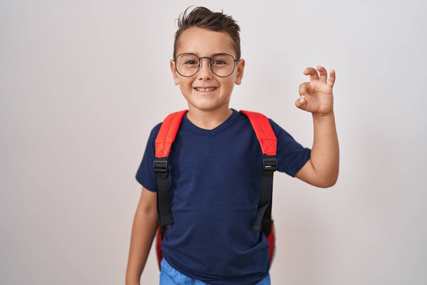Küçük İspanyol çocuk gözlük takıyor ve öğrenci çantası takıyor. Parmaklarıyla imza atıyor, gülümsüyor. Mükemmel bir sembol.  - Fotoğraf, Görsel