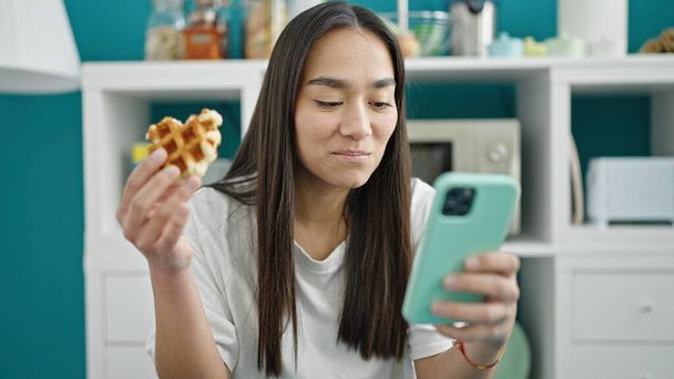 Genç ve güzel İspanyol kadın waffle yiyor. Yemek odasında akıllı telefon kullanıyor. - Fotoğraf, Görsel