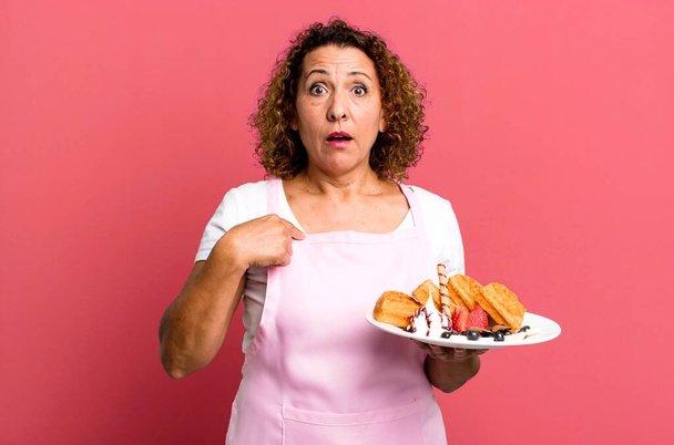 pretty middle age woman looking shocked and surprised with mouth wide open, pointing to self. home made waffles concept - Foto, Imagen
