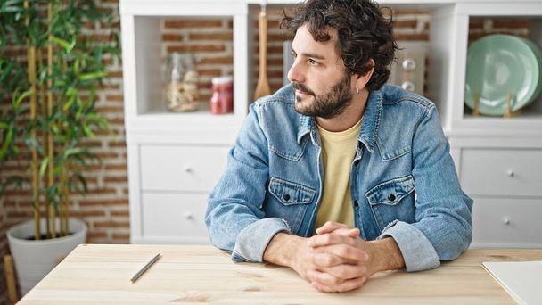Young hispanic man sitting on table with relaxed expression at dinning room - 写真・画像