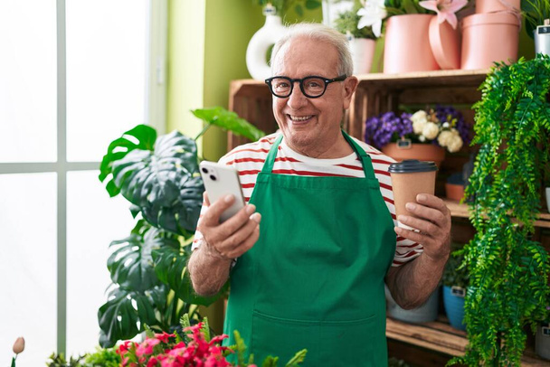 Middle age grey-haired man florist using smartphone drinking coffee at flower shop - Photo, Image