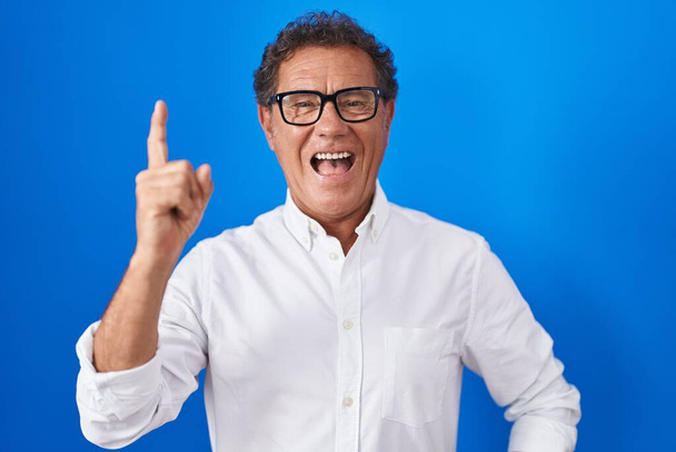 Латиноамериканец средних лет, стоящий на синем фоне, указывая пальцем на успешную идею. Взволнован и счастлив. номер один.  - Фото, изображение