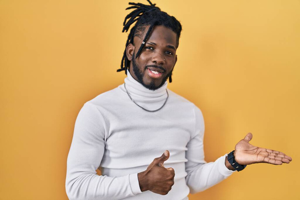 African man with dreadlocks wearing turtleneck sweater over yellow background showing palm hand and doing ok gesture with thumbs up, smiling happy and cheerful  - Photo, Image