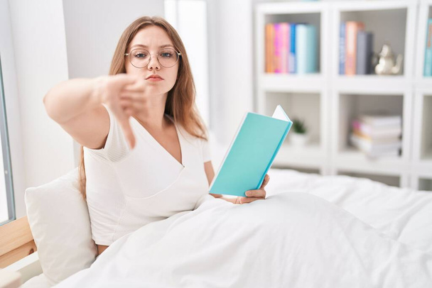 Młoda biała kobieta siedząca w domu na łóżku i czytająca książkę z rozgniewaną twarzą, negatywna oznaka niechęci do opuszczenia kciuków, koncepcja odrzucenia  - Zdjęcie, obraz