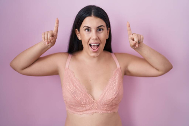 Young hispanic woman wearing pink bra smiling amazed and surprised and pointing up with fingers and raised arms.  - Photo, Image