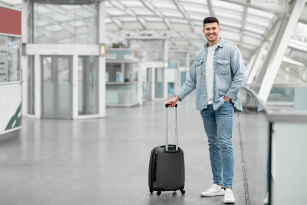 Utazási ajánlat a Globetrotternek. Férfi utazó pózol bőrönd poggyász álló vigyorog a kamera a repülőtéren beltéri. Teljes hosszúságú felvétel fiatal utasokról, akik a repülésre várnak. Fénymásolási tér - Fotó, kép