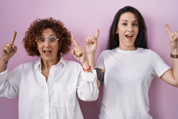 Madre e hija hispanas vistiendo camiseta blanca casual sobre fondo rosa sonriendo asombradas y sorprendidas y señalando con los dedos y los brazos levantados.  - Foto, imagen