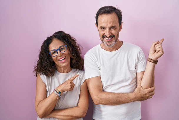 Middle age hispanic couple together over pink background with a big smile on face, pointing with hand finger to the side looking at the camera.  - Фото, зображення