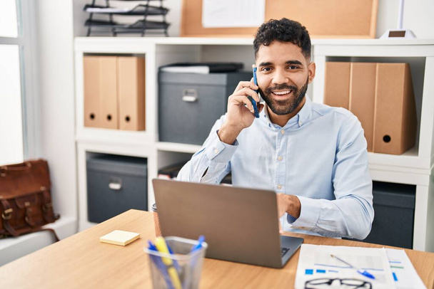 Jonge Spaanse man zakenman met behulp van laptop praten op de smartphone op kantoor - Foto, afbeelding