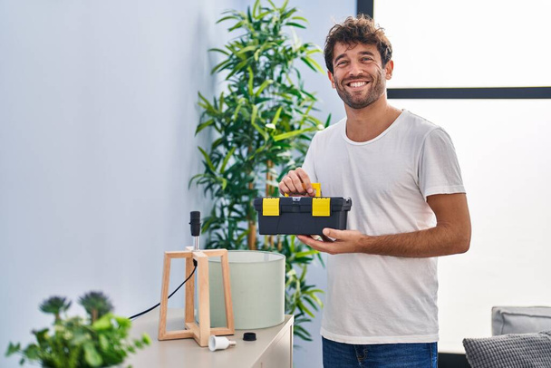Junger Mann lächelt zuversichtlich und hält Werkzeugkiste zu Hause - Foto, Bild