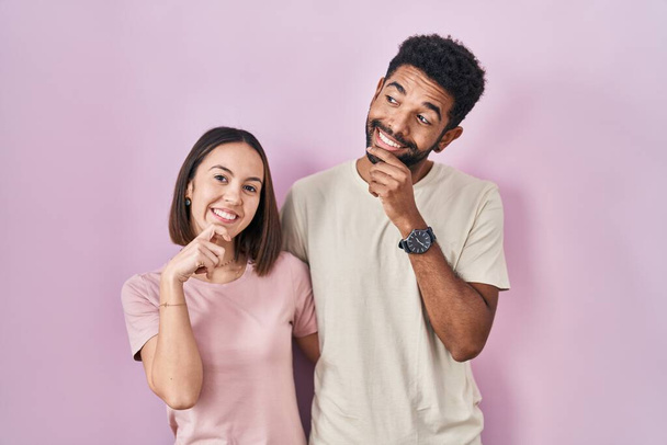 Young hispanic couple together over pink background with hand on chin thinking about question, pensive expression. smiling and thoughtful face. doubt concept.  - Photo, Image
