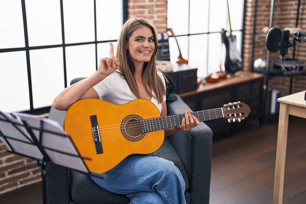 Giovane bella donna che suona la chitarra classica in studio di musica sorpreso con un'idea o una domanda che punta il dito con la faccia felice, numero uno  - Foto, immagini