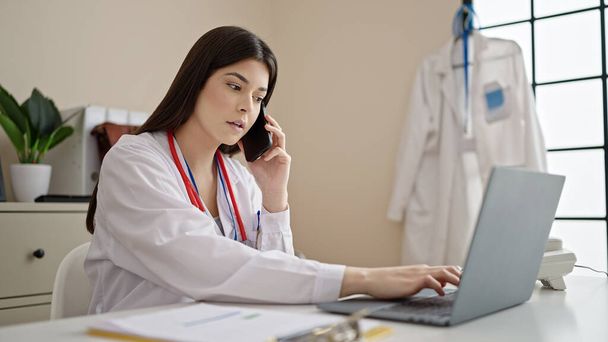 Mladá krásná hispánská žena lékař pomocí notebooku mluvit na smartphone na klinice - Fotografie, Obrázek
