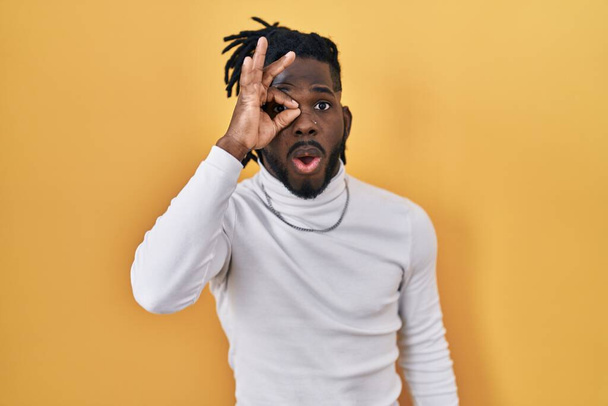 African man with dreadlocks wearing turtleneck sweater over yellow background doing ok gesture shocked with surprised face, eye looking through fingers. unbelieving expression.  - Foto, imagen