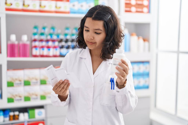Young woman pharmacist holding pills bottle reading prescription at pharmacy - Photo, Image