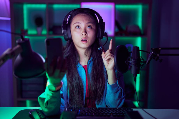 Молода азіатка грає у відеоігри зі смартфоном здивована і здивована, дивлячись вгору і вказуючи пальцями і піднятими руками
.  - Фото, зображення